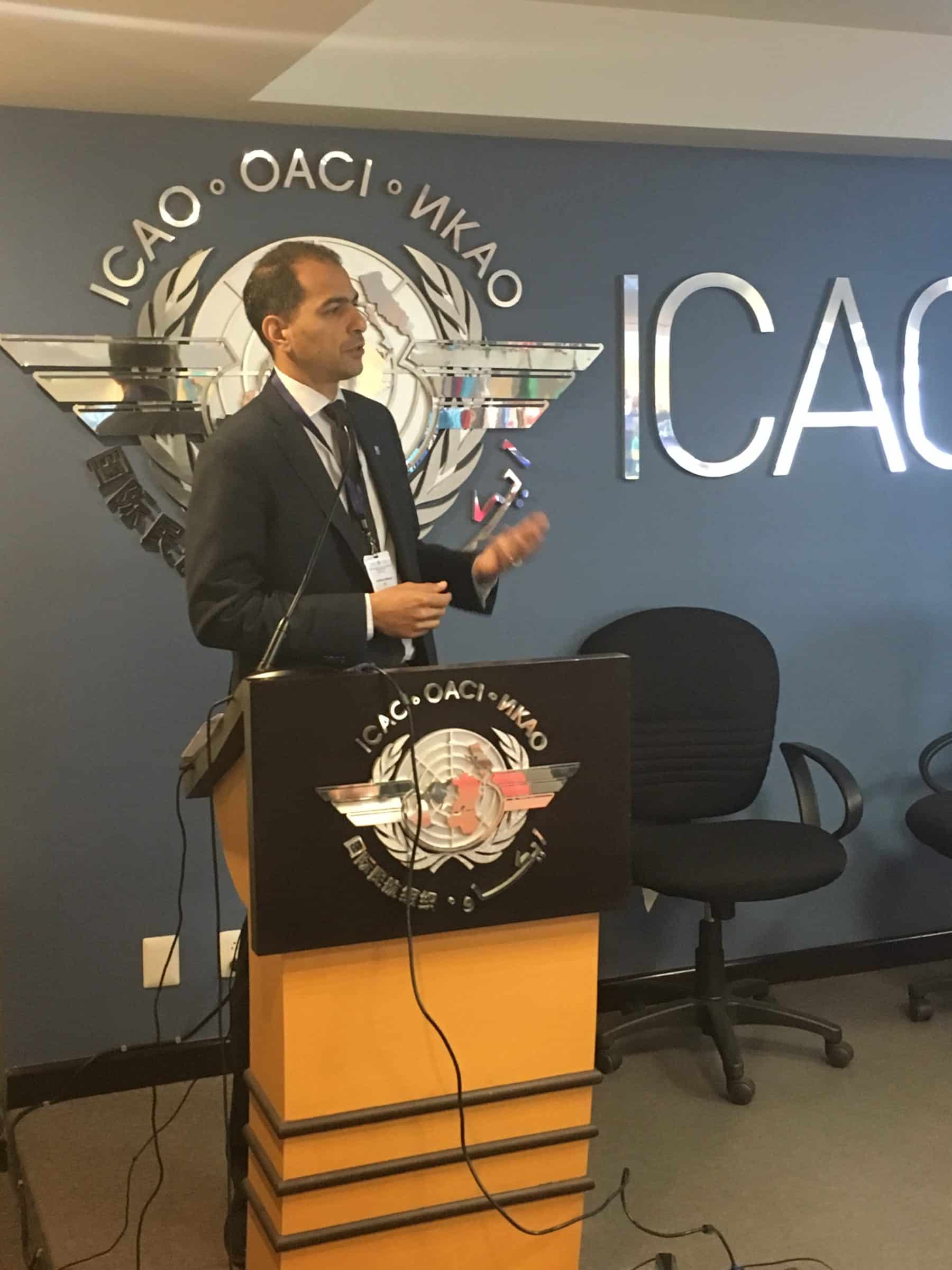 A man in a suit stands at a podium giving a presentation in front of an ICAO logo on the wall, with empty chairs beside him.