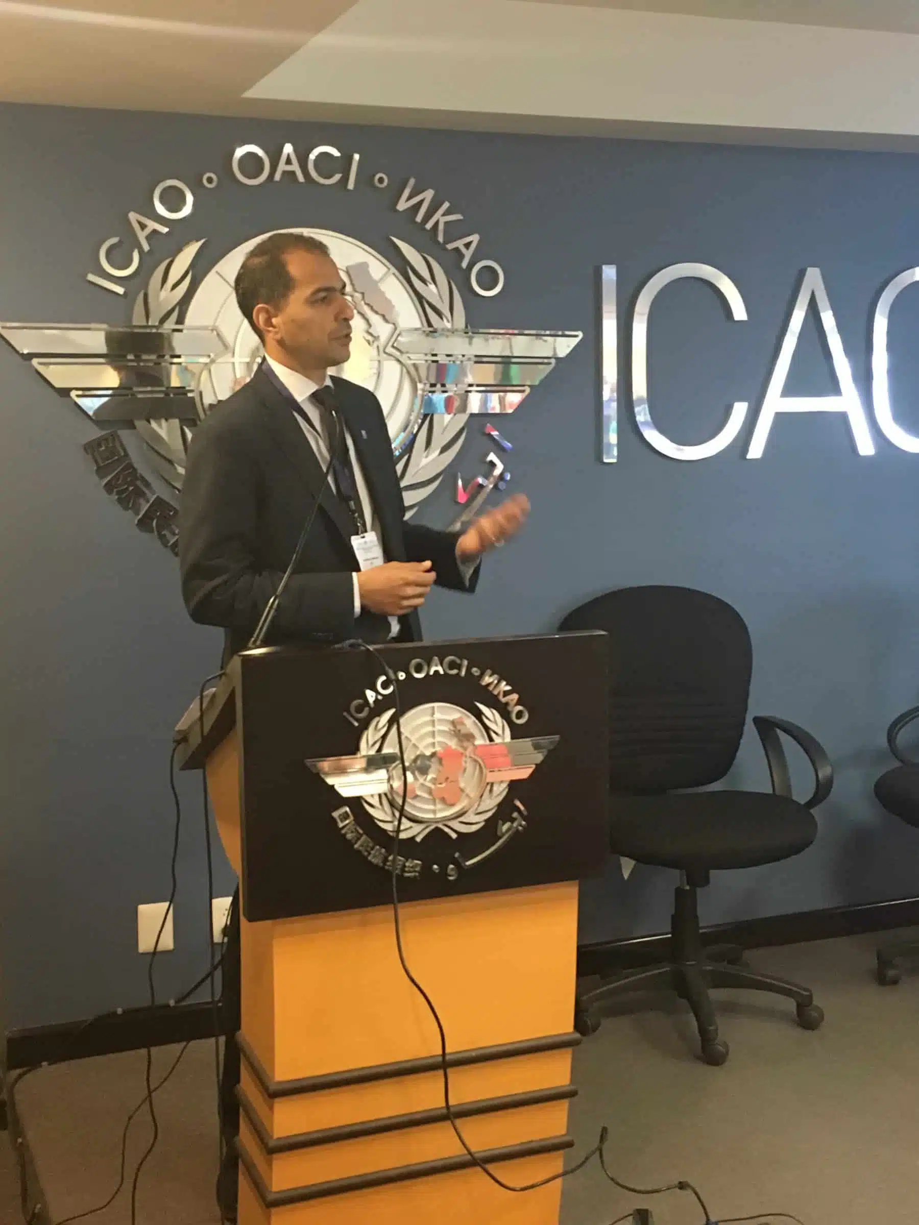 A man in a suit stands at a podium giving a presentation in front of an ICAO logo on the wall, with empty chairs beside him.