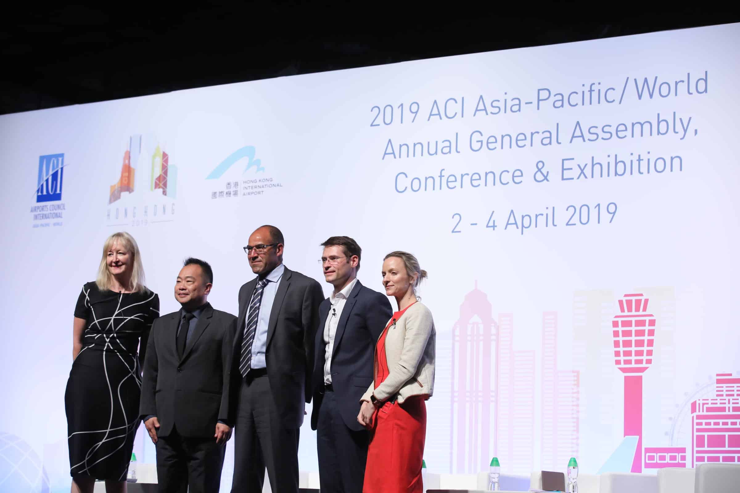 Five people in business attire stand on stage in front of a large screen displaying 2019 ACI Asia-Pacific/World Annual General Assembly, Conference & Exhibition, 2-4 April 2019 with event logos and cityscape graphics.