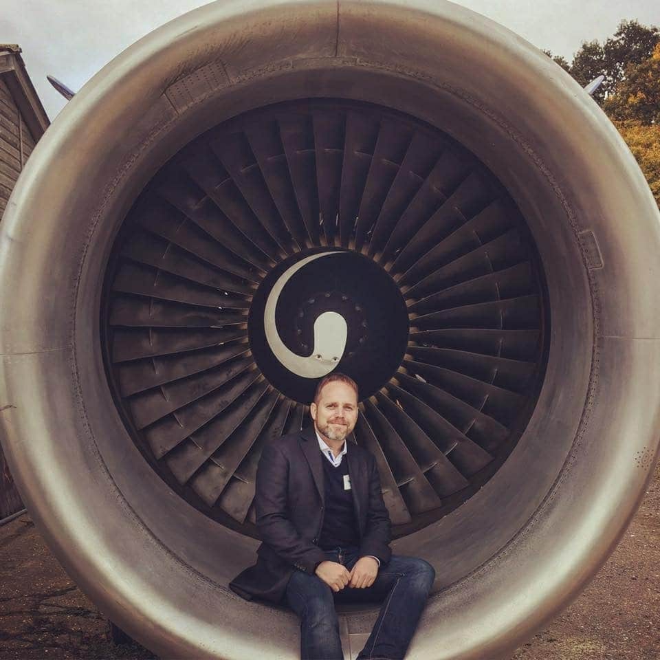 A man in a suit sits inside the hollow center of a large airplane engine turbine, smiling at the camera. The fan blades form a circular pattern behind him.