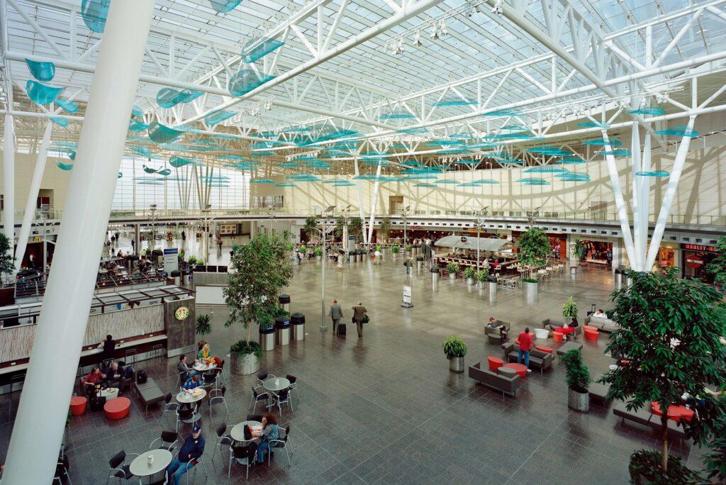 Spacious airport terminal with high glass ceilings, scattered seating, indoor trees, several people walking or sitting, shops and food stalls along the sides, and blue decorative elements hanging from the ceiling.