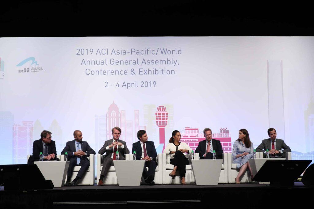 Seven people in business attire sit on a stage panel during the 2019 ACI Asia-Pacific/World Annual General Assembly, Conference & Exhibition. A large event banner is displayed behind them with the event details.