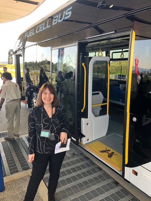 A woman stands smiling in front of an open door of a modern fuel cell bus. People are gathered around and some greenery is visible in the background.