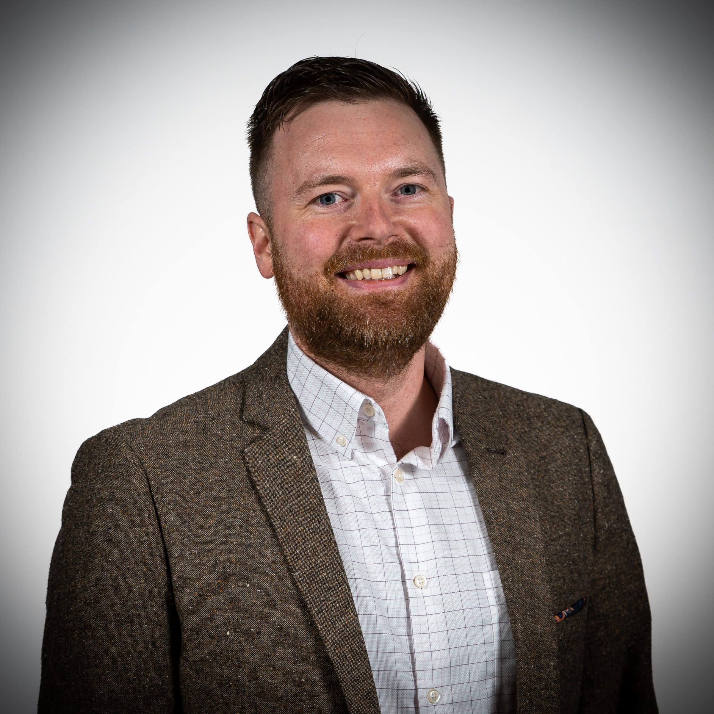A man with short brown hair and a beard, wearing a brown tweed blazer and a white checkered shirt, smiles in front of a plain white background.