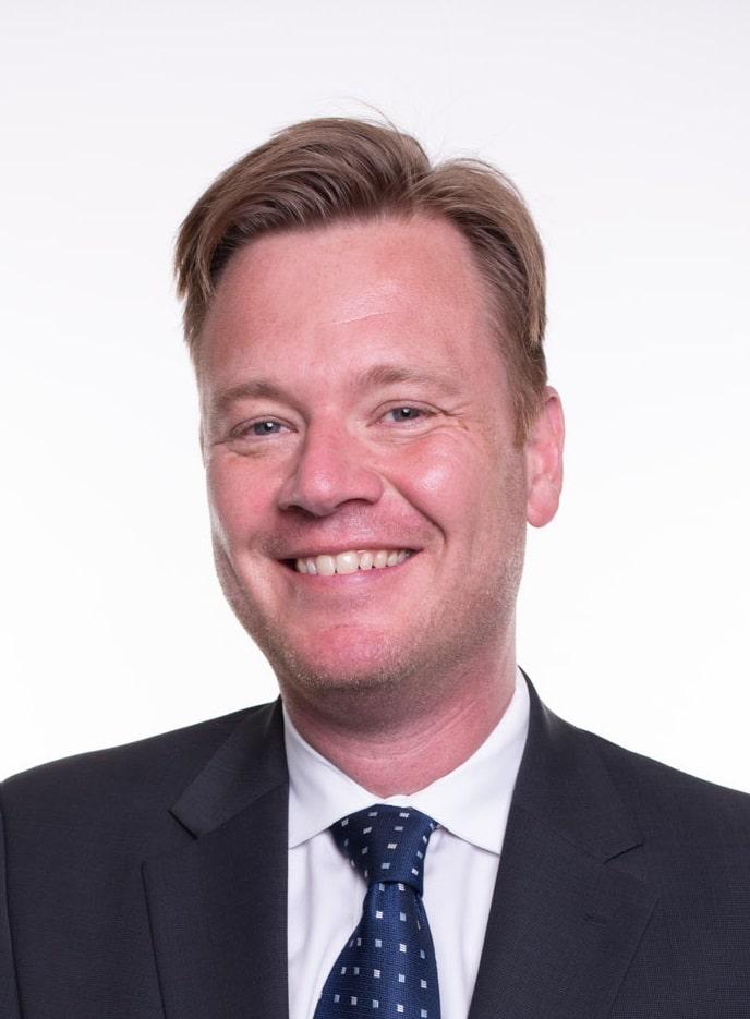 A man in a dark suit and blue polka-dot tie smiles at the camera against a plain white background. He has light brown hair and a fair complexion.
