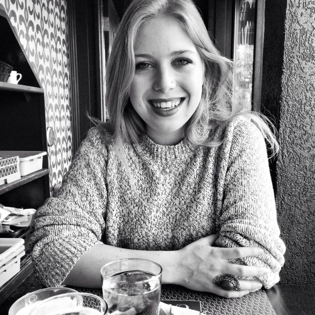 A young woman with long hair sits at a table, smiling warmly. She wears a knit sweater and has her arms folded. On the table are a glass of water and a cup. The photo is in black and white.