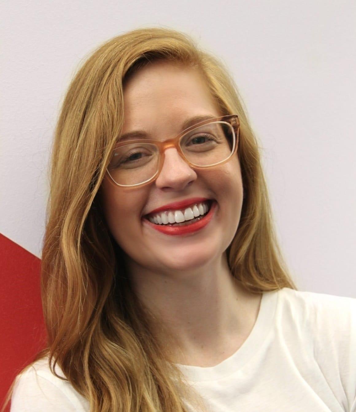 A young woman with long, wavy blonde hair and glasses smiles broadly. She is wearing a white shirt and standing against a white and red background.