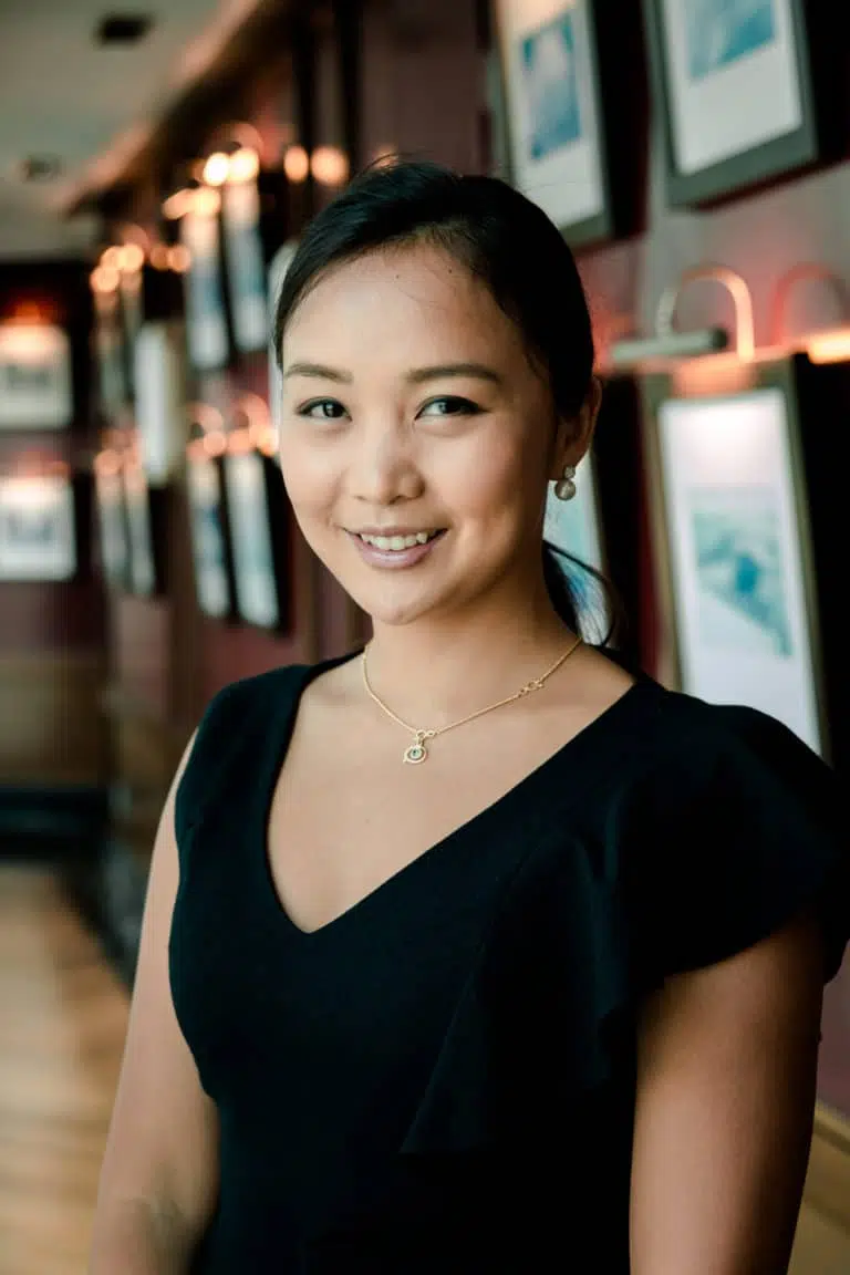 A woman with dark hair tied back, wearing a black dress and gold necklace, stands smiling in a warmly lit hallway with framed pictures on the wall behind her.