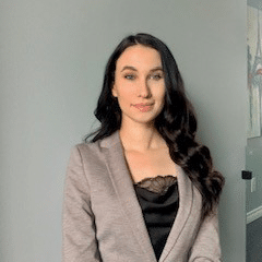 A woman with long dark hair wearing a gray blazer and a black top stands in front of a light gray wall, looking at the camera with a slight smile.