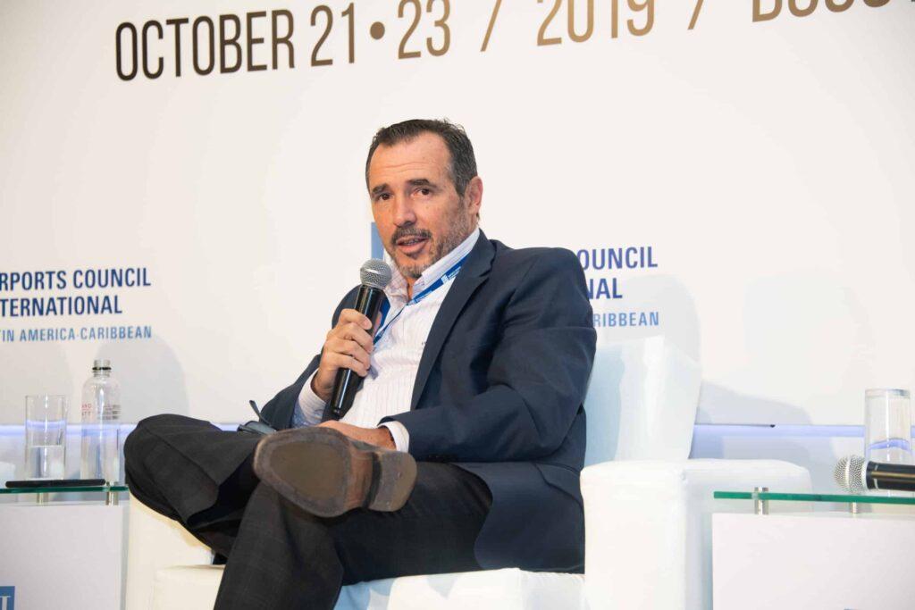 A man in a dark suit sits on a white chair, holding a microphone and speaking at a conference. The background displays event information, including the dates October 21-23, 2019, and the venue name.