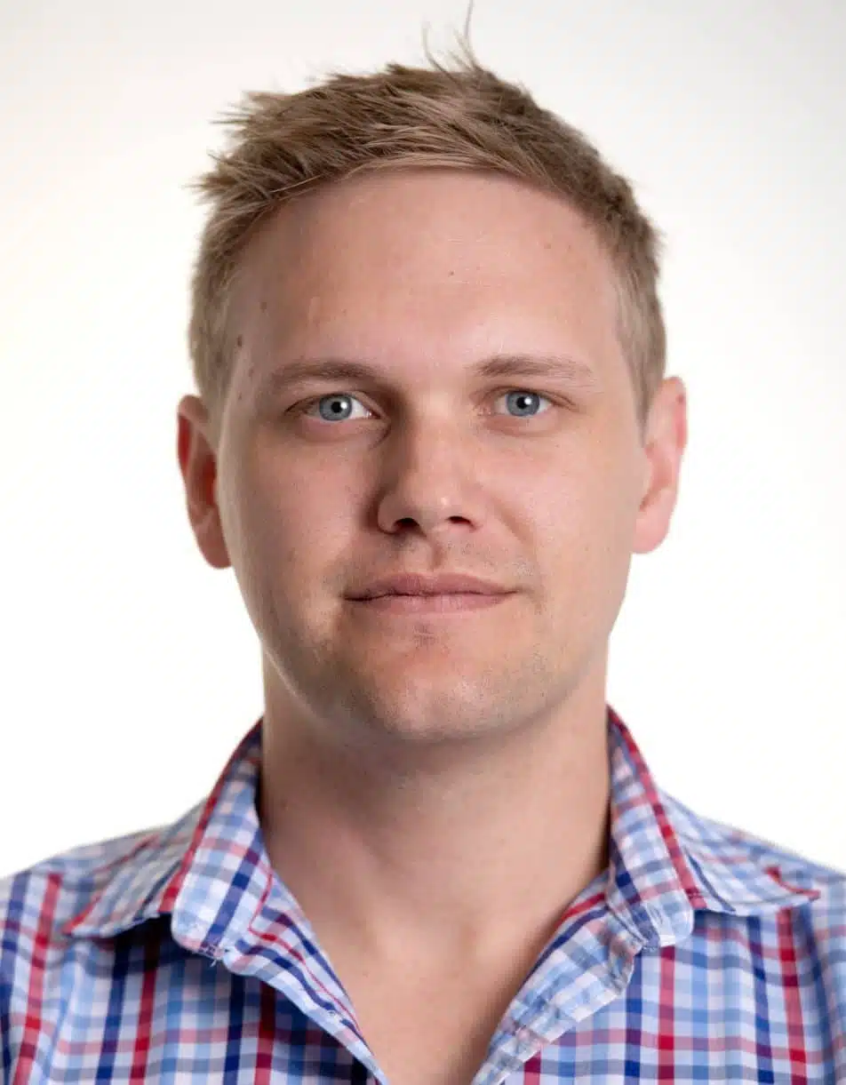 A man with short blond hair and blue eyes wearing a blue, red, and white plaid shirt, looking directly at the camera against a white background.