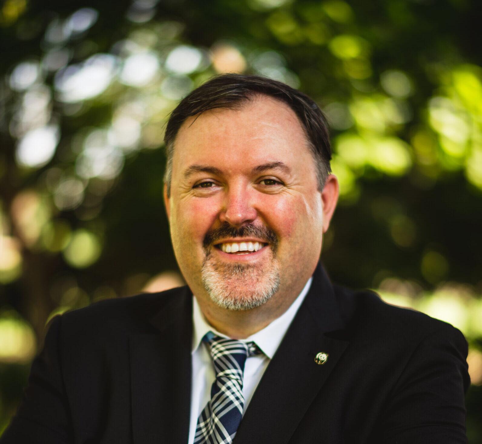 A man in a suit and plaid tie smiles outdoors, standing in front of trees and greenery with sunlight filtering through the leaves.