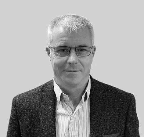 A man with short gray hair, glasses, and a neutral expression is wearing a striped shirt and a dark textured blazer, posing against a plain light gray background.