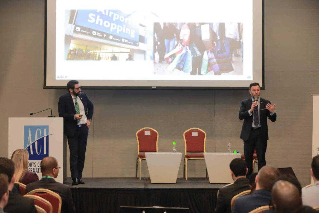 Two men in suits speak on stage at a conference with a screen behind them displaying images related to airport shopping. They stand beside two empty chairs and small tables with water bottles. Audience members watch attentively.