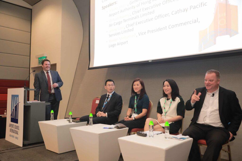 Four panelists sit on stage with microphones while a speaker stands at a podium. A projected slide with speaker names is displayed behind them. All are dressed in business attire at a professional event.