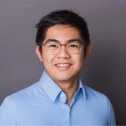 A young man with short dark hair, wearing glasses and a light blue button-up shirt, smiles in front of a plain gray background.