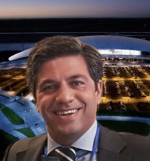 A smiling man in a suit and tie is in the foreground, with a large, modern, well-lit building and a parking lot filled with cars at dusk in the background.