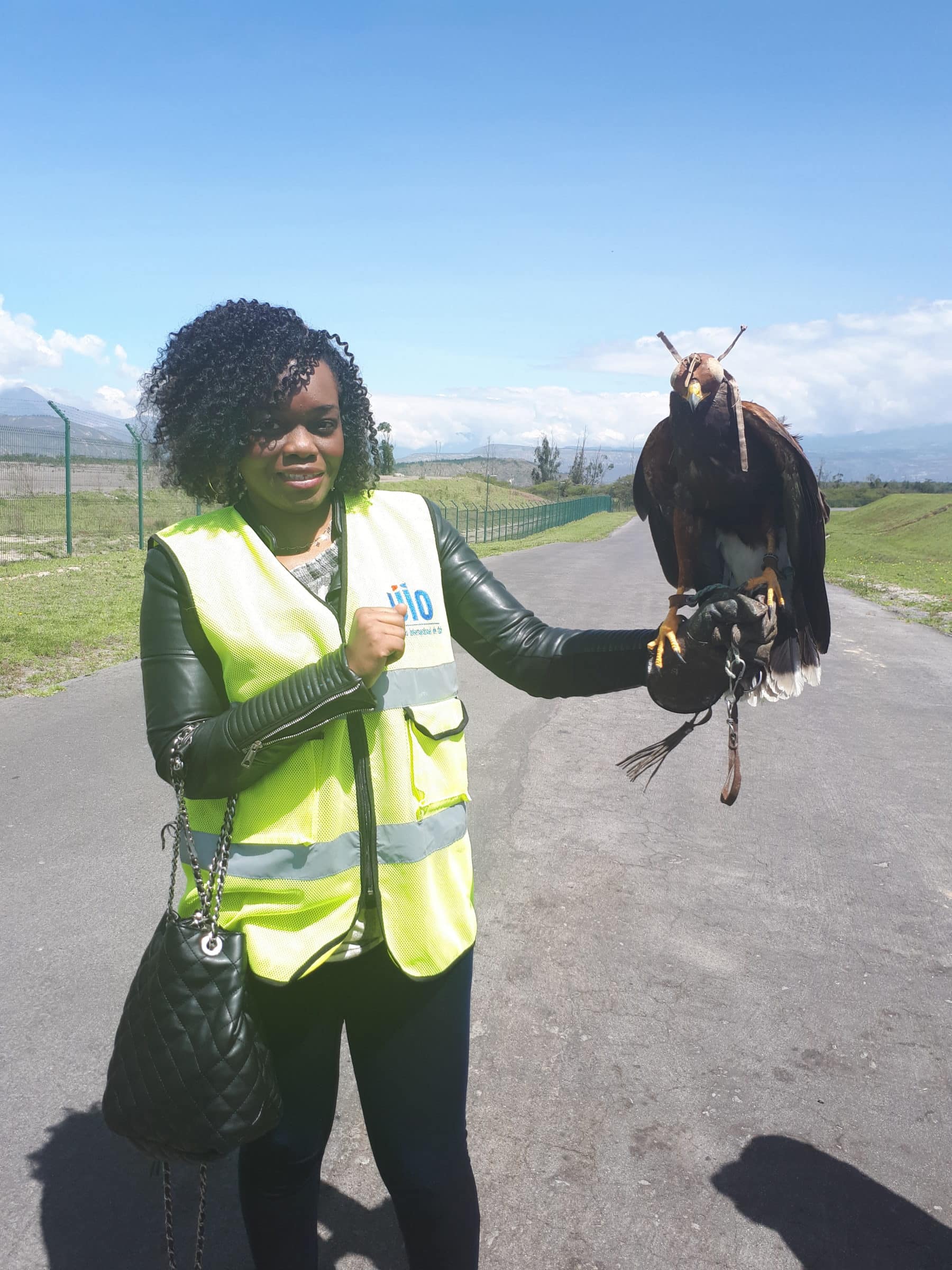 A woman wearing a yellow safety vest and black gloves stands on a paved road, holding a large brown bird of prey on her gloved hand. She smiles at the camera; mountains and blue sky are in the background.