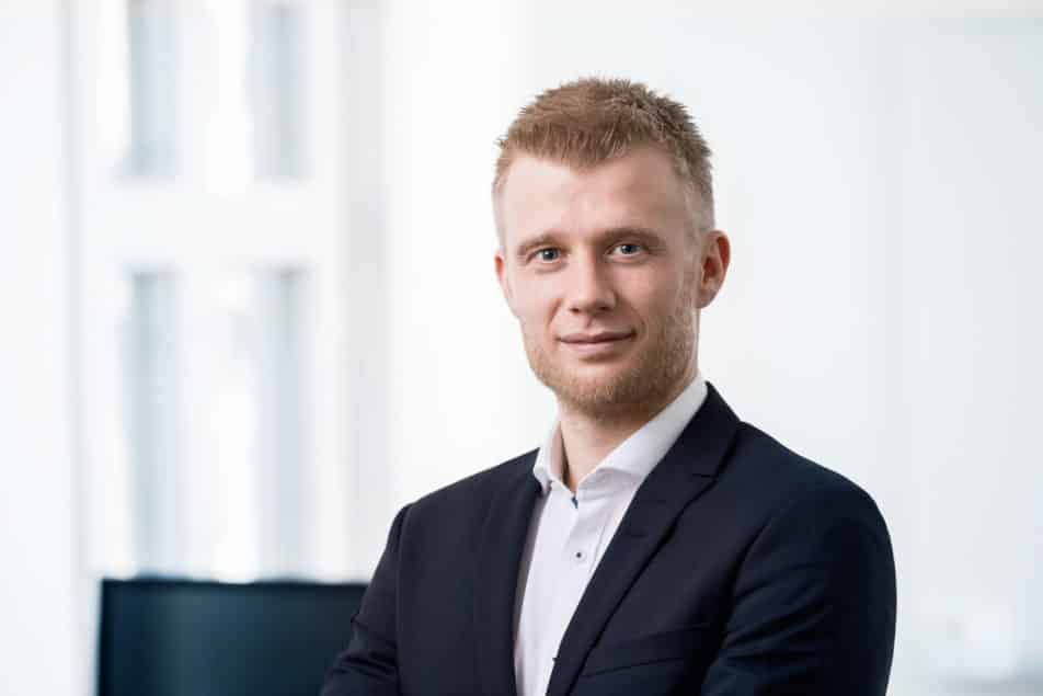 A young man with short blond hair and a trimmed beard, wearing a dark suit jacket and white shirt, stands confidently indoors with a slight smile. The background is bright and out of focus.