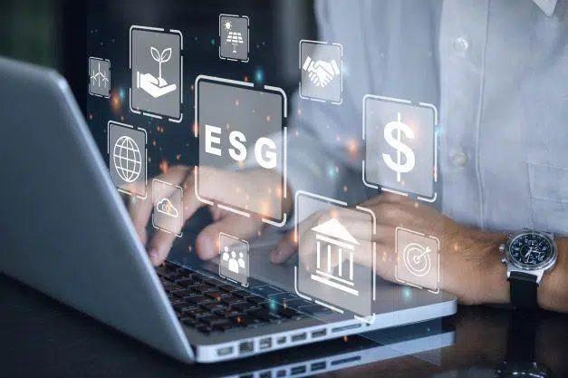 A person types on a laptop with floating digital icons representing ESG (Environmental, Social, Governance) concepts, financial symbols, and sustainability elements appearing in the foreground.