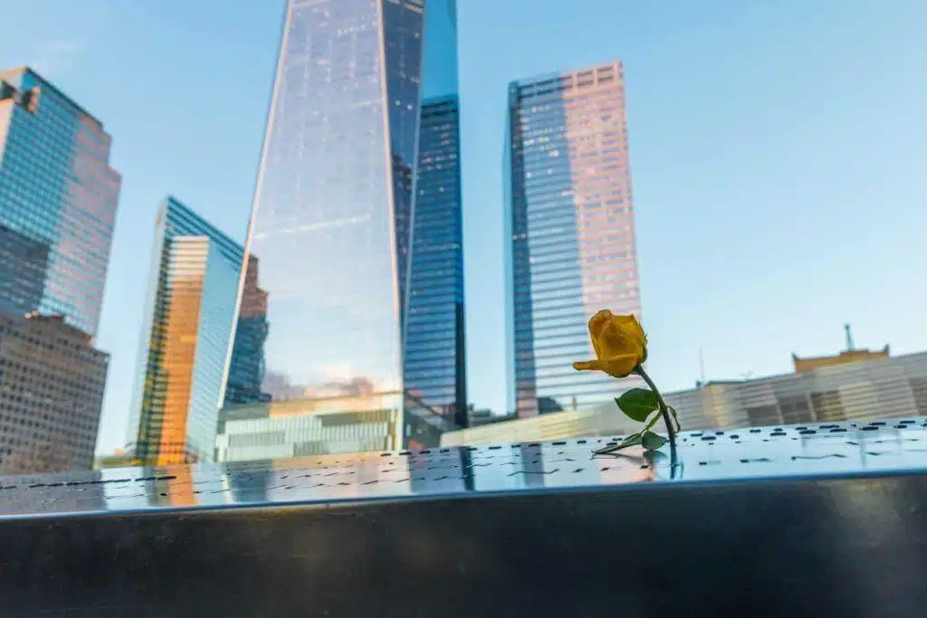 A single yellow rose rests on the engraved names at the 9/11 Memorial in New York City, with tall glass skyscrapers reflecting the sky in the background.