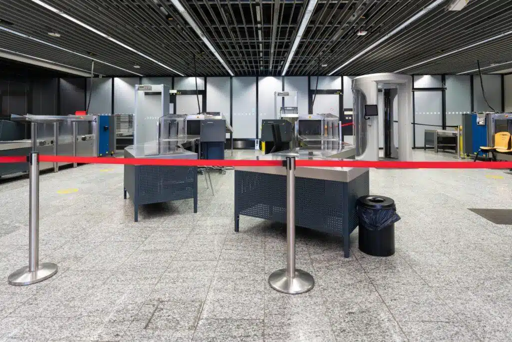 An empty airport security checkpoint with metal detectors, baggage scanners, gray flooring, a red barrier tape, and no people present. The area looks clean and organized under bright overhead lighting.