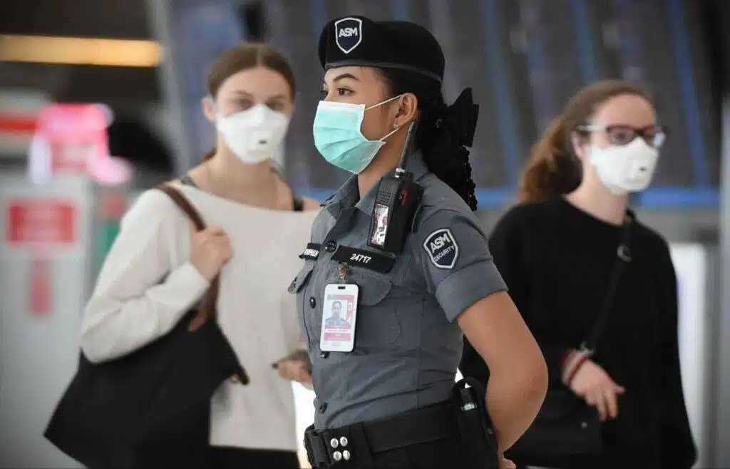 A security officer wearing a uniform and face mask stands in front of two people, also in face masks, at what appears to be an indoor public space or transportation hub.