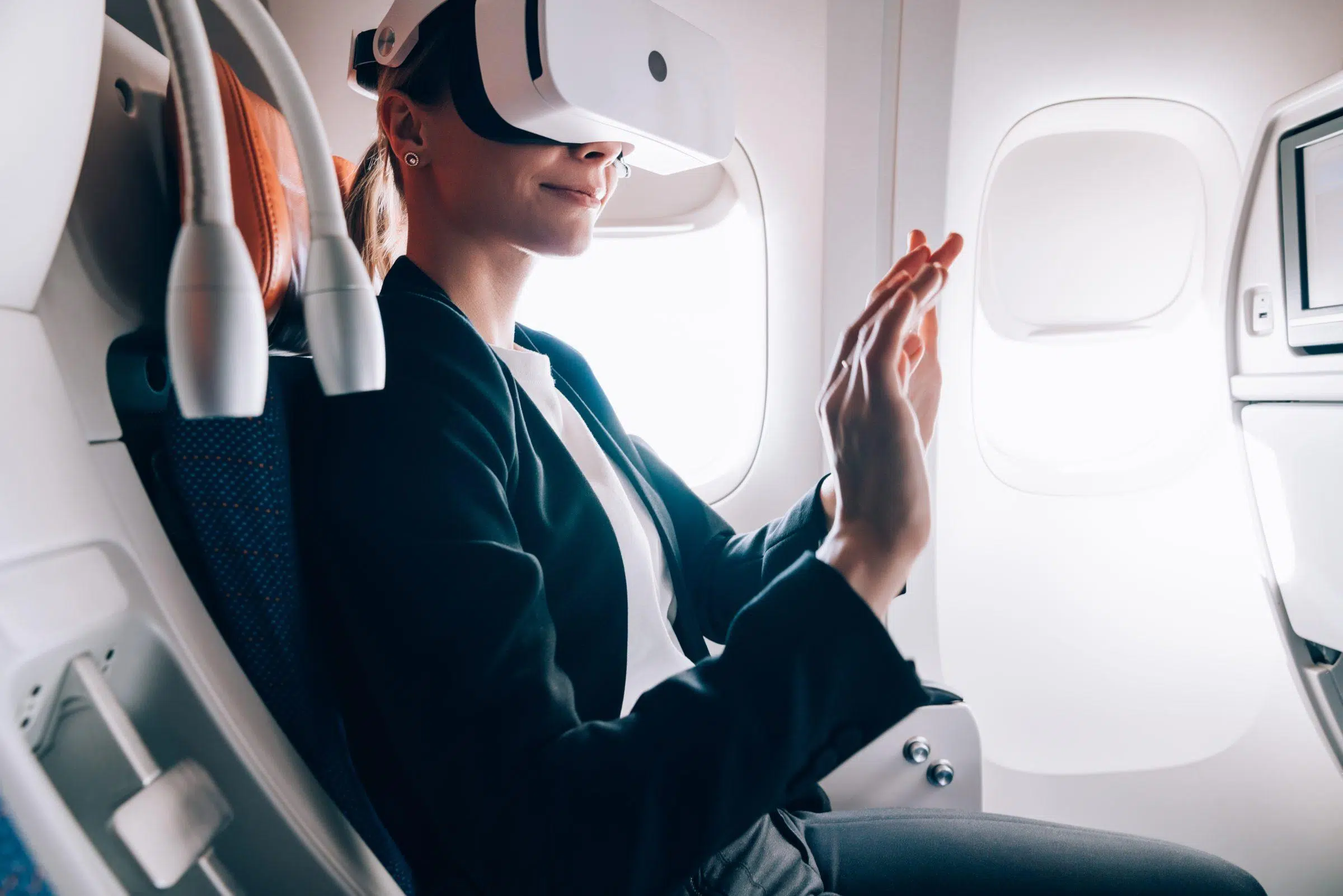 A woman sits in an airplane seat wearing a virtual reality headset, with her hands raised in front of her as if interacting with a virtual environment. Sunlight streams through the nearby window.