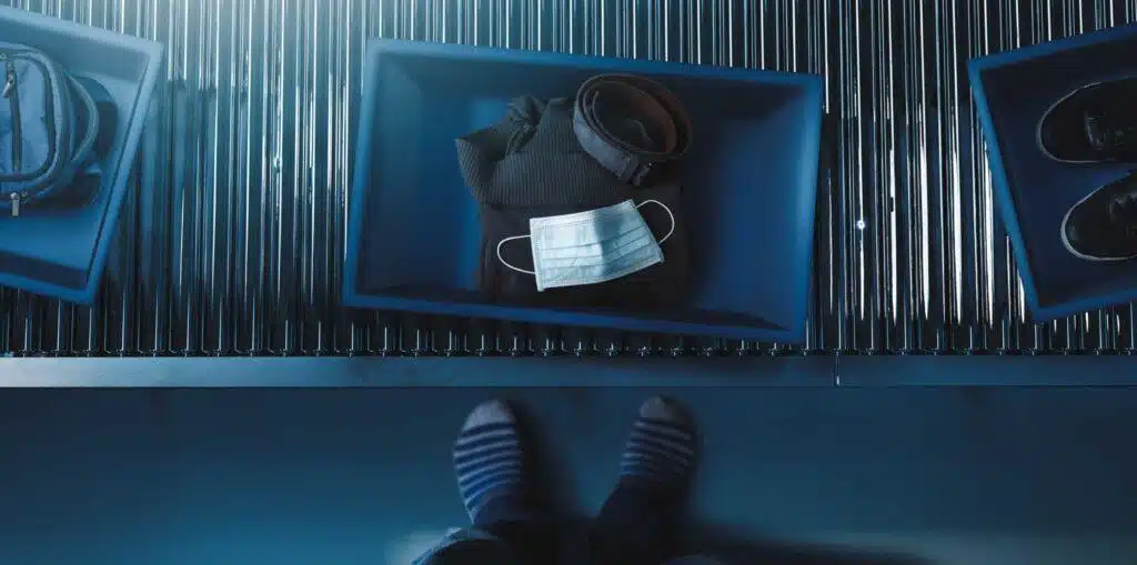 A blue tray on an airport security conveyor belt holds a folded jacket, a belt, and a disposable face mask. The tray is surrounded by other bins, and a person’s feet are visible at the bottom of the image.