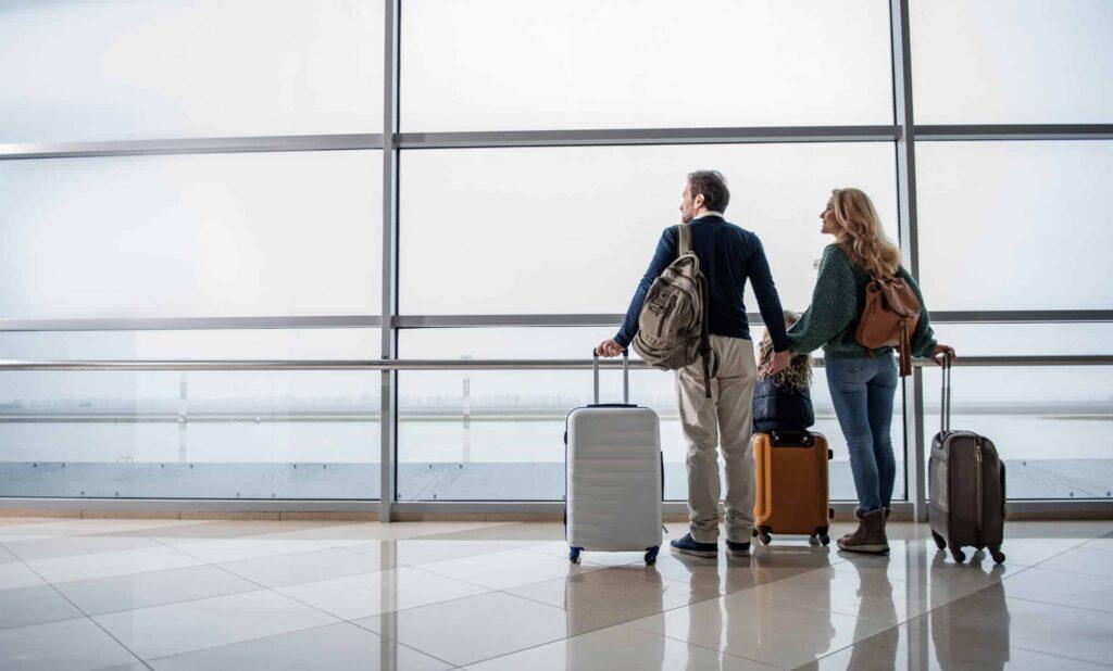 A man and woman stand side by side in an airport terminal, holding hands and looking out large windows. They each have a suitcase and backpacks, ready for travel.