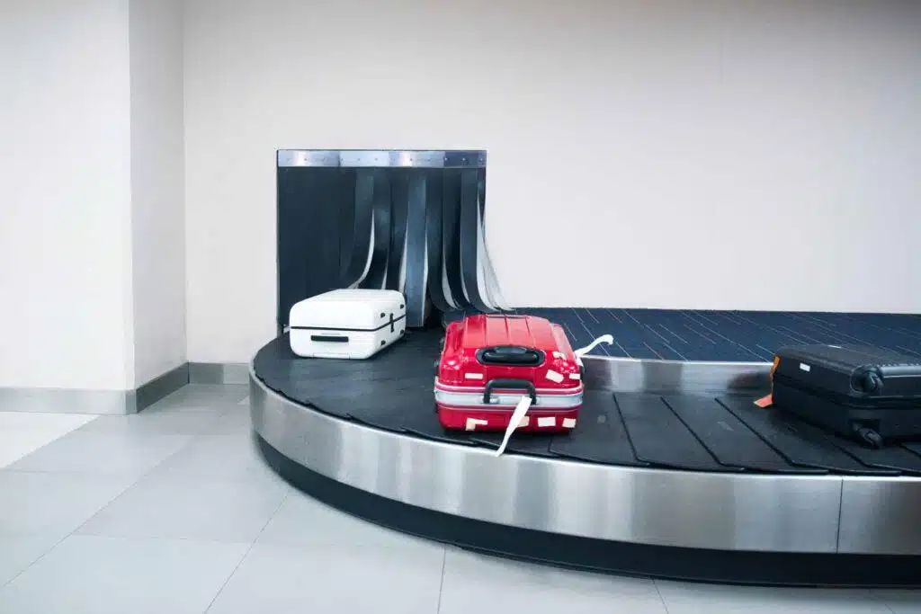 Three suitcases—white, red, and black—on an airport baggage carousel against a plain wall, with the red suitcase standing out in the center.