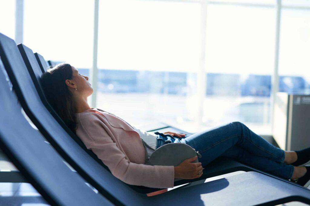A woman reclines and rests on a lounge chair in a brightly lit airport terminal, holding a small gray bag on her lap, with large windows and blurry airplanes visible in the background.