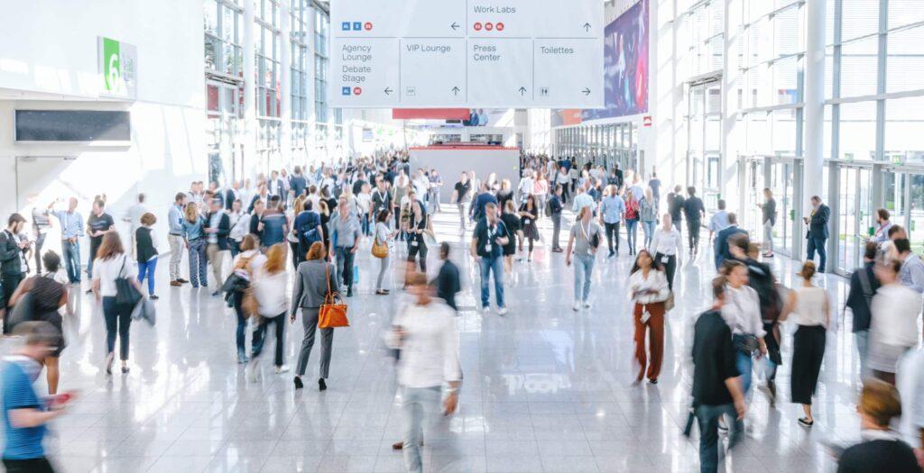 A large crowd of people walks through a bright, spacious convention center hallway with glass walls and high ceilings. Overhead signs point to various areas such as the agency lounge, VIP lounge, and press center.