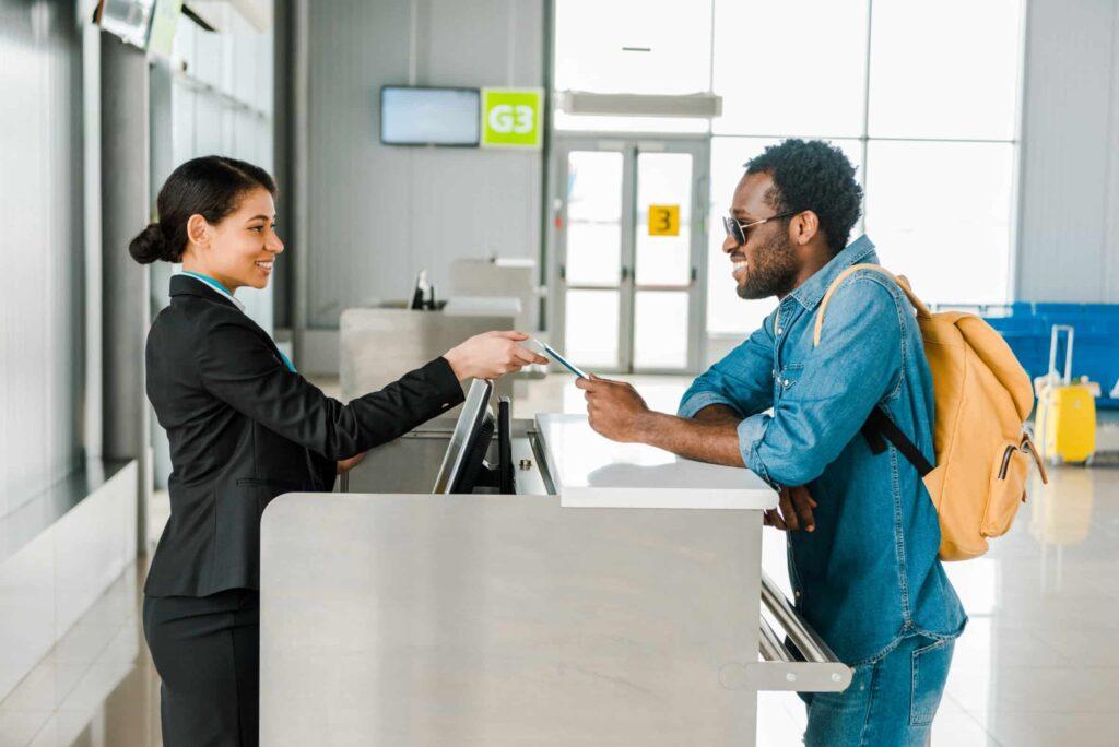 A smiling airline staff member hands a boarding pass to a man wearing sunglasses and a backpack at an airport check-in counter. The terminal is bright with signs and luggage visible in the background.