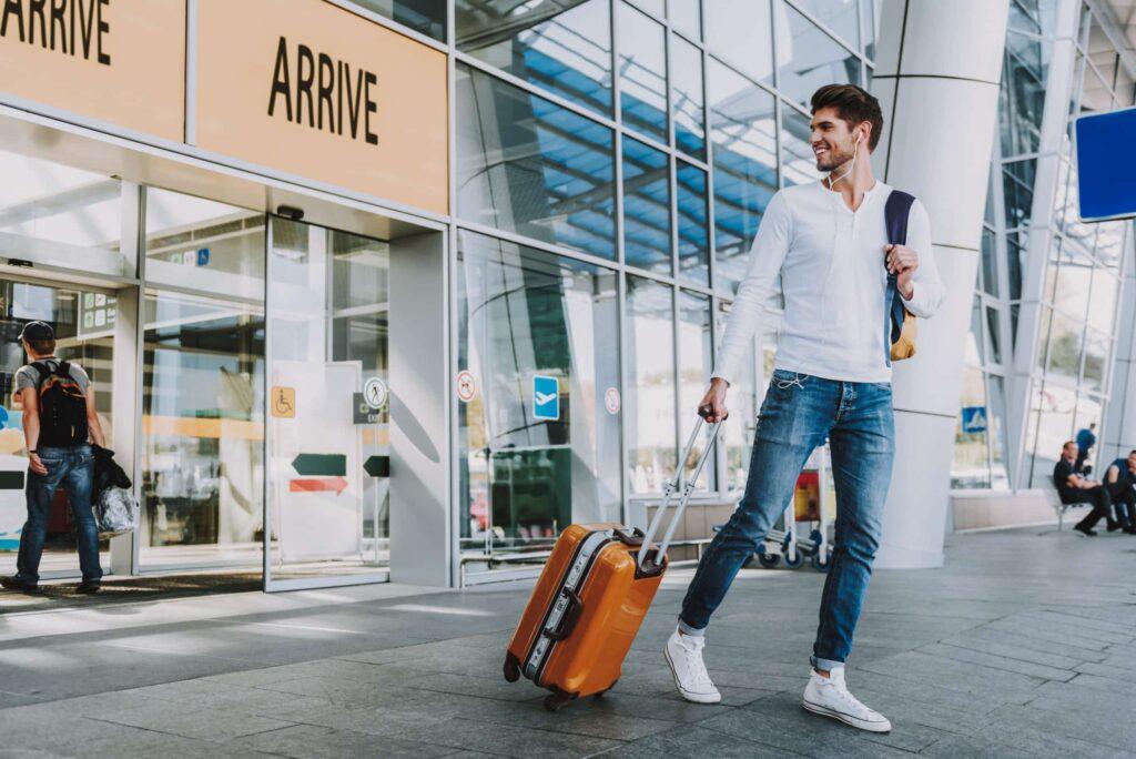 A young man wearing jeans and a white shirt smiles while pulling an orange suitcase outside an airport arrival terminal, with glass walls and automatic doors in the background.