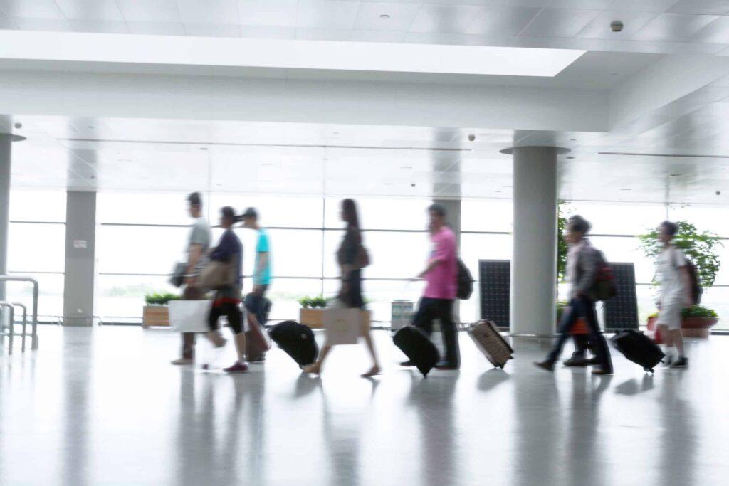 A group of people walk through a bright, modern airport terminal, each pulling rolling suitcases. The image is slightly blurred, emphasizing their movement. Large windows and potted plants are visible in the background.