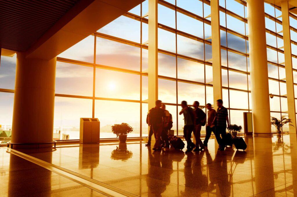 A group of people with suitcases walk through a spacious airport terminal with large windows, as sunlight streams in, creating reflections on the shiny floor.