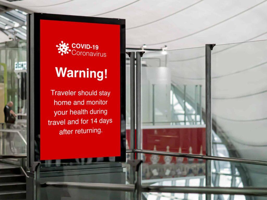 A digital sign in an airport displays a red COVID-19 warning, advising travelers to stay home and monitor their health during travel and for 14 days after returning. The background shows a modern, glass-walled terminal.