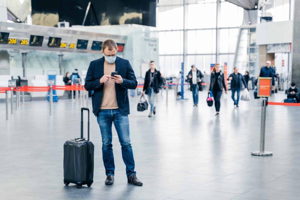 A man wearing a face mask stands in an airport terminal, looking at his phone with a suitcase by his side. Other travelers are walking in the background near check-in counters.