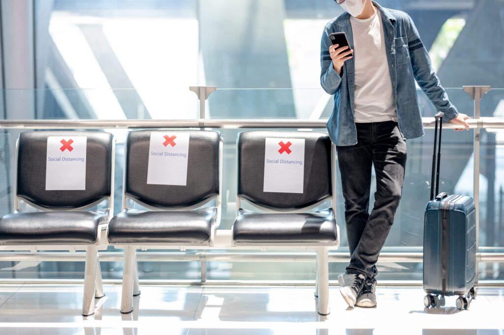 A person wearing a mask stands in an airport with a suitcase, holding a phone. Three empty seats beside them have signs with red Xs and Social Distancing written on them.