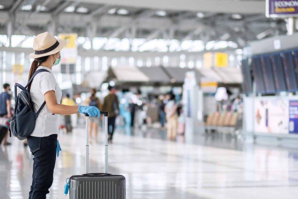 A woman wearing a hat, face mask, gloves, and backpack stands in an airport terminal with a suitcase, looking at a digital flight information board. Other travelers and airport signs are visible in the background.