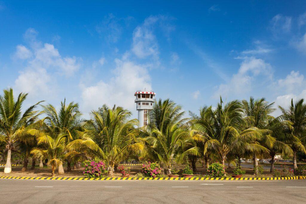 A control tower rises above lush palm trees and flowering bushes under a blue sky with scattered clouds, viewed from across an empty paved road with yellow-and-black painted curb.