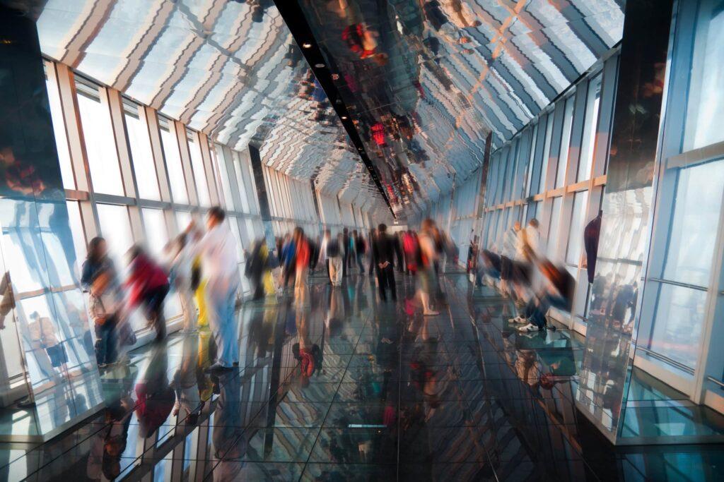 A group of people, blurred by motion, walk and stand along a glass-floor observation deck inside a modern skyscraper with large windows and angled glass ceilings reflecting the scene.
