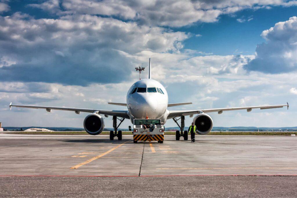 A large commercial airplane is parked on an airport tarmac under a partly cloudy sky, with a ground crew member standing nearby in a high-visibility vest.