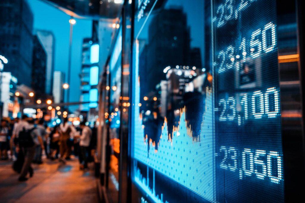 A digital stock market display shows fluctuating numbers and graphs at night, reflecting city lights and blurred pedestrians walking along a busy urban street.