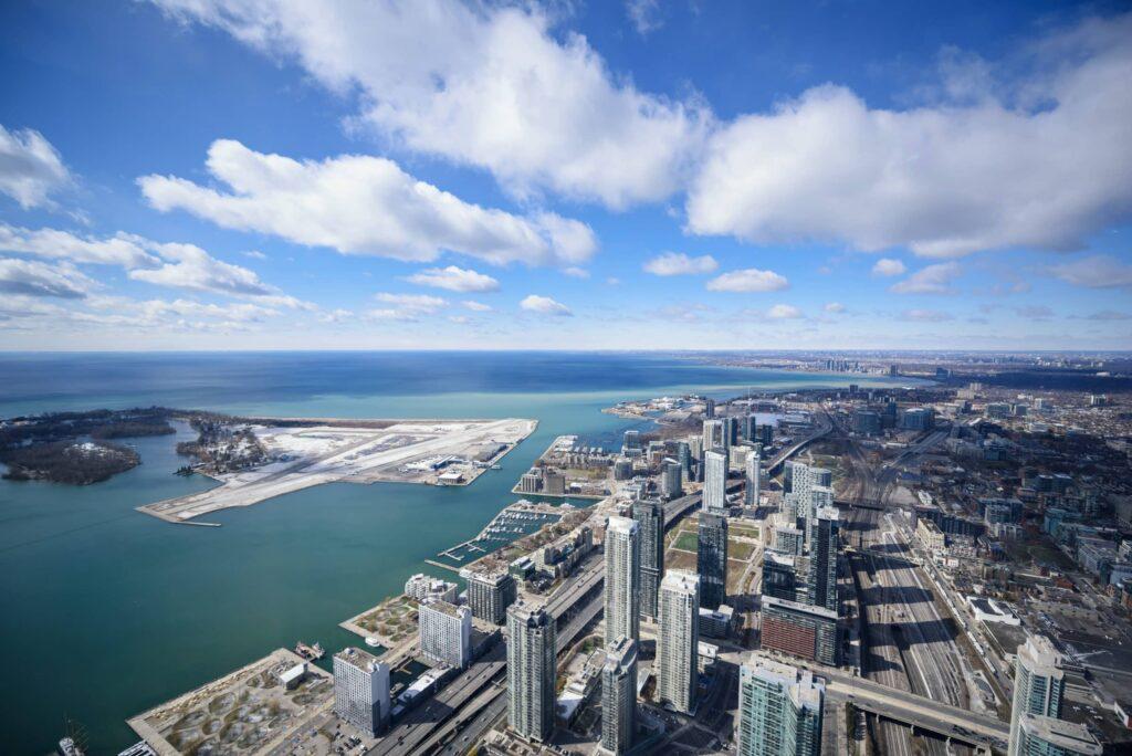 Aerial view of a city skyline with tall buildings near a waterfront, a small airport on a peninsula, and a large body of water under a blue sky with scattered clouds.
