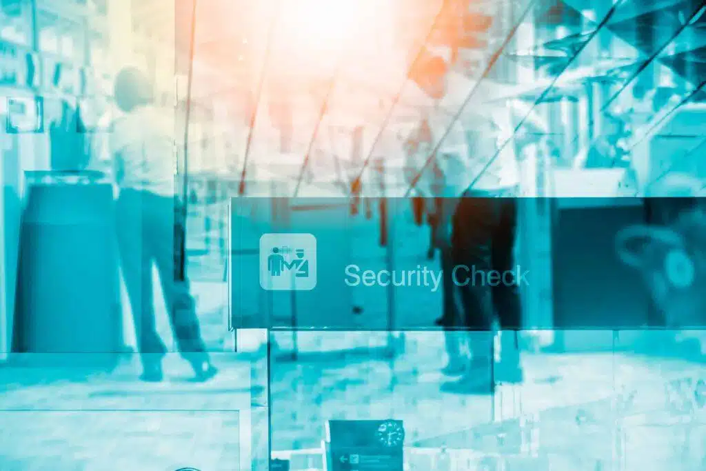 Blurry image of people standing near a glass wall with a sign that reads “Security Check,” suggesting an airport or secure facility entrance. Sunlight creates a blue and orange overlay effect.