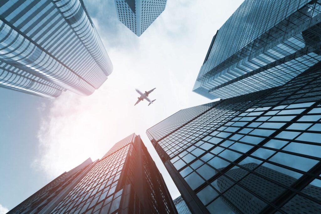 A commercial airplane flies overhead, framed by tall, modern glass skyscrapers against a bright sky, viewed from ground level looking up.
