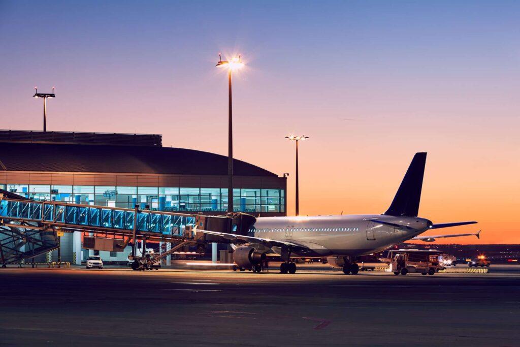 A commercial airplane is parked at an airport gate with a jet bridge connected, as the sun sets in the background. The terminal building is lit up, and airport vehicles are nearby.