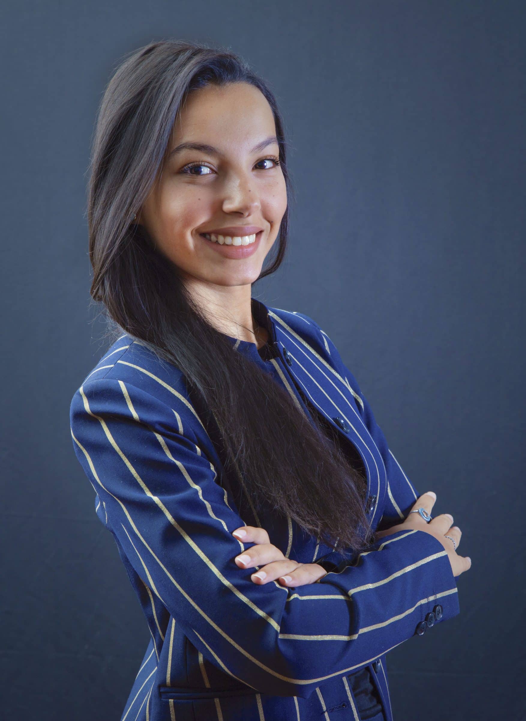 A woman with long dark hair, wearing a blue pinstripe blazer, stands against a plain dark background. She is smiling confidently with her arms crossed.